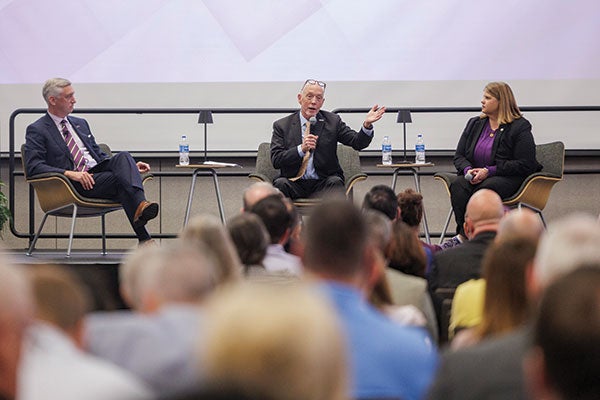 A crowd listens to two men and a woman in suits talking.