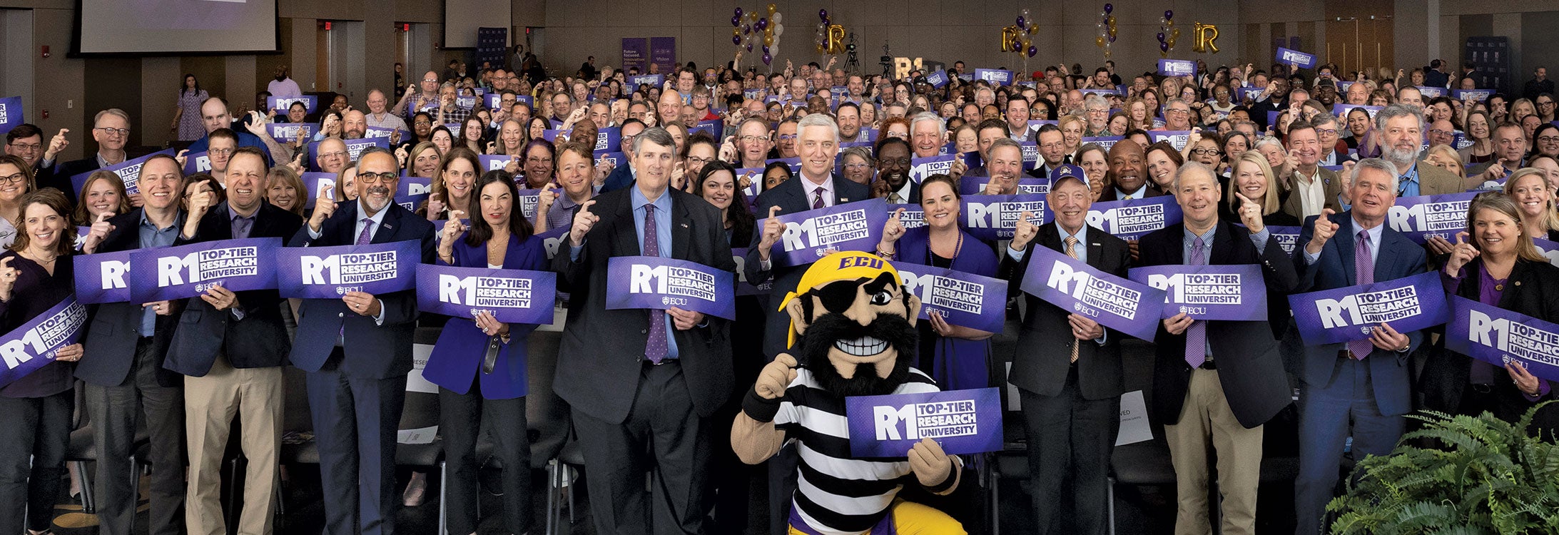 A Pirate mascot kneels in front of a crowd holding R1 signs