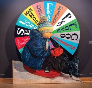 A young boy in winter outdoor clothing sits on a sled. A wheel is behind him and is divided into multicolored wedges. Each wedge contains a word related to growing up.