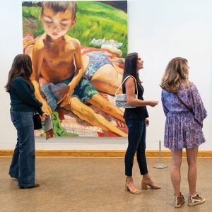 Three patrons look at a large painting of a young boy wearing a bathing suit. He is seated on a blanket in some grass.