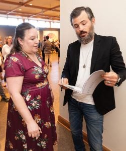 Scott Avett looks at a booklet he's holding while speaking with a woman in a magenta floral dress.