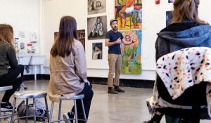 Scott Avett stands in front of a wall of artwork and speaks to art students seated on metal stools.