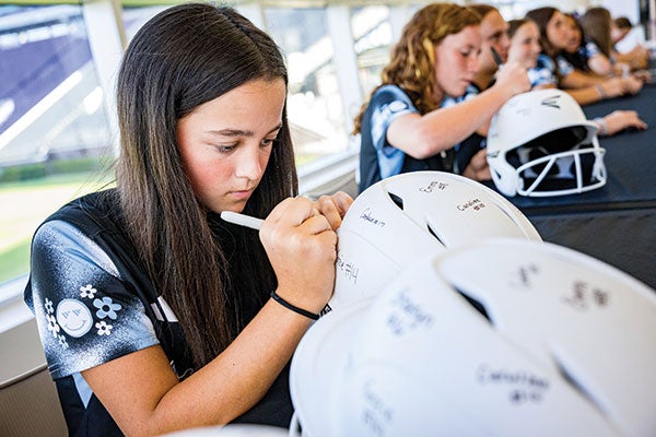 A young girl signing her name on a baseball helmet, showcasing her personal touch and enthusiasm for the sport.