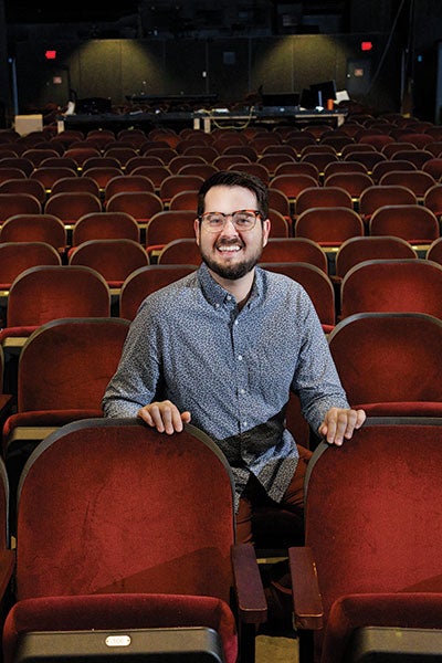 A man seated in an auditorium, surrounded by vibrant red seats, focused on the stage ahead. 