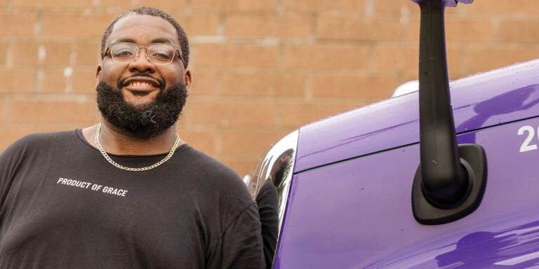 A bearded man stands beside a purple truck, showcasing a casual outdoor scene.