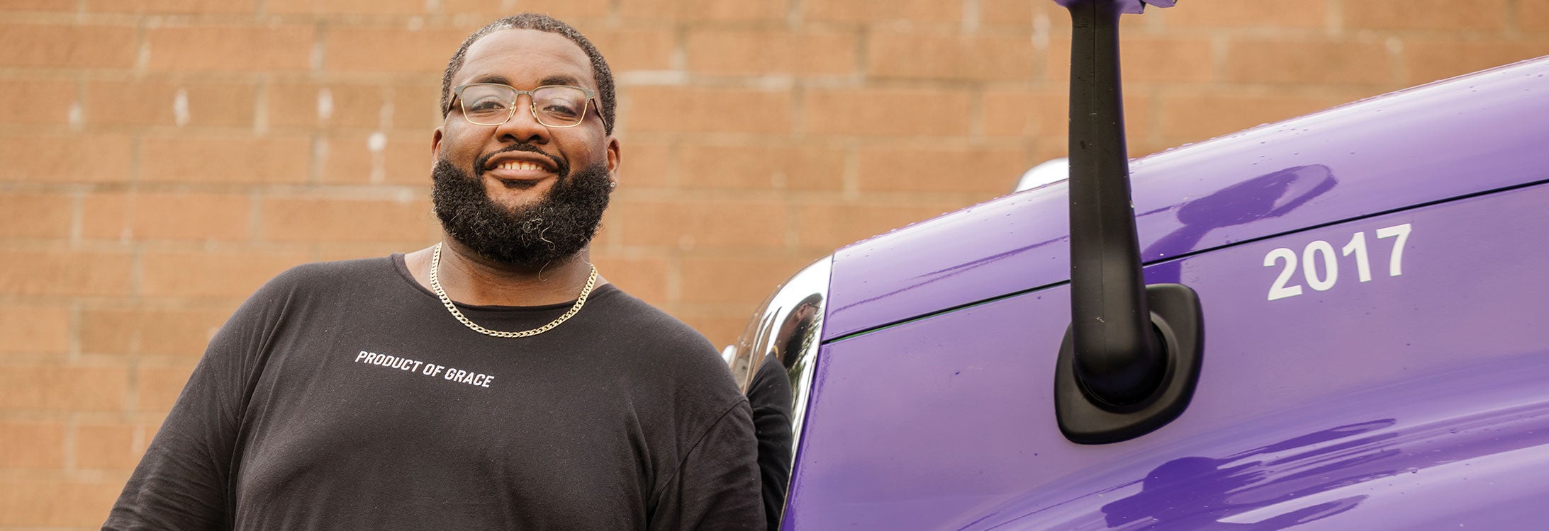 A bearded man stands beside a purple truck, showcasing a casual outdoor scene.