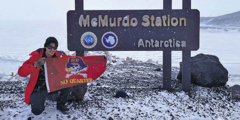 A researcher sits in front of a sign for McMurdo Station in Anatarctica