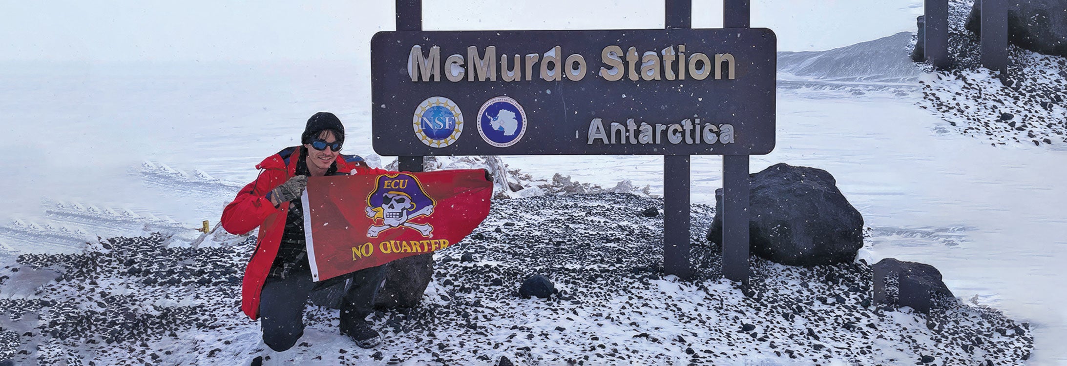 A researcher sits in front of a sign for McMurdo Station in Anatarctica