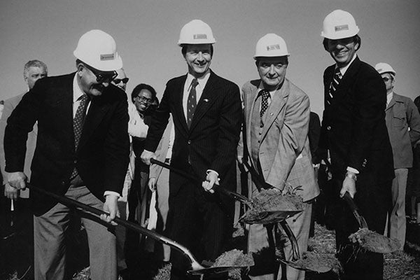 Four men in suits and hard hats stand together, each holding a shovel, ready for a groundbreaking ceremony.