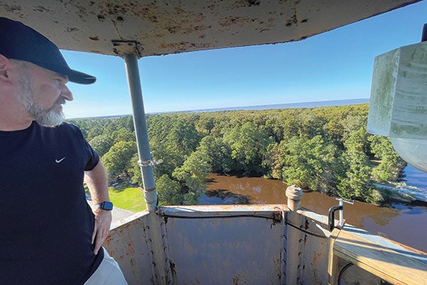 A man stands atop a tower, gazing out over a serene body of water, with a clear sky in the background.