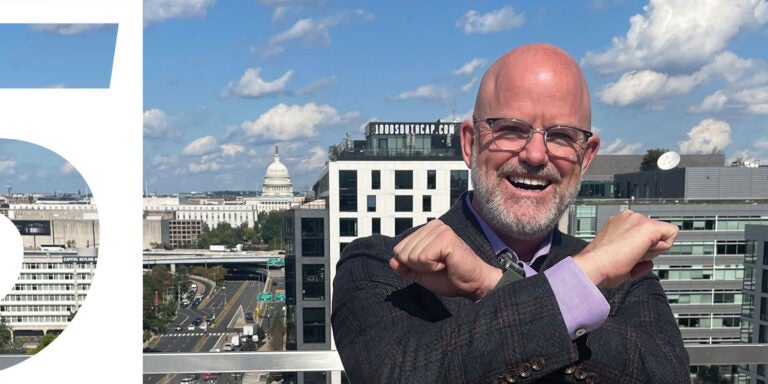 A man with glasses and a beard stands on a balcony, overlooking a vibrant city skyline in the background.