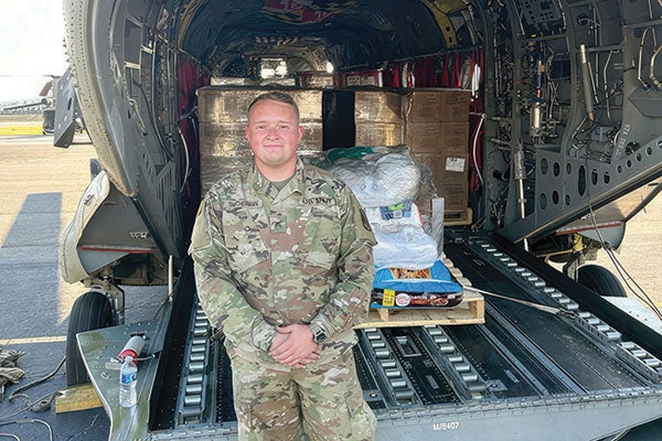 A man in military uniform stands proudly before a large military cargo plane, showcasing his dedication and service.